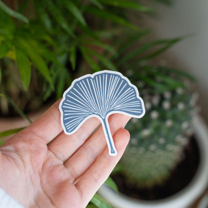 Hand holding a blue ginkgo leaf stickers in front of a houseplant